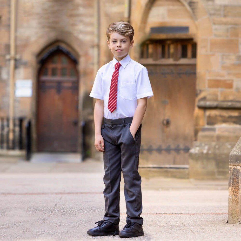 
                  
                    A young boy wearing a white school shirt, red tie, and grey trousers stands confidently outside of his school entrance door.
                  
                